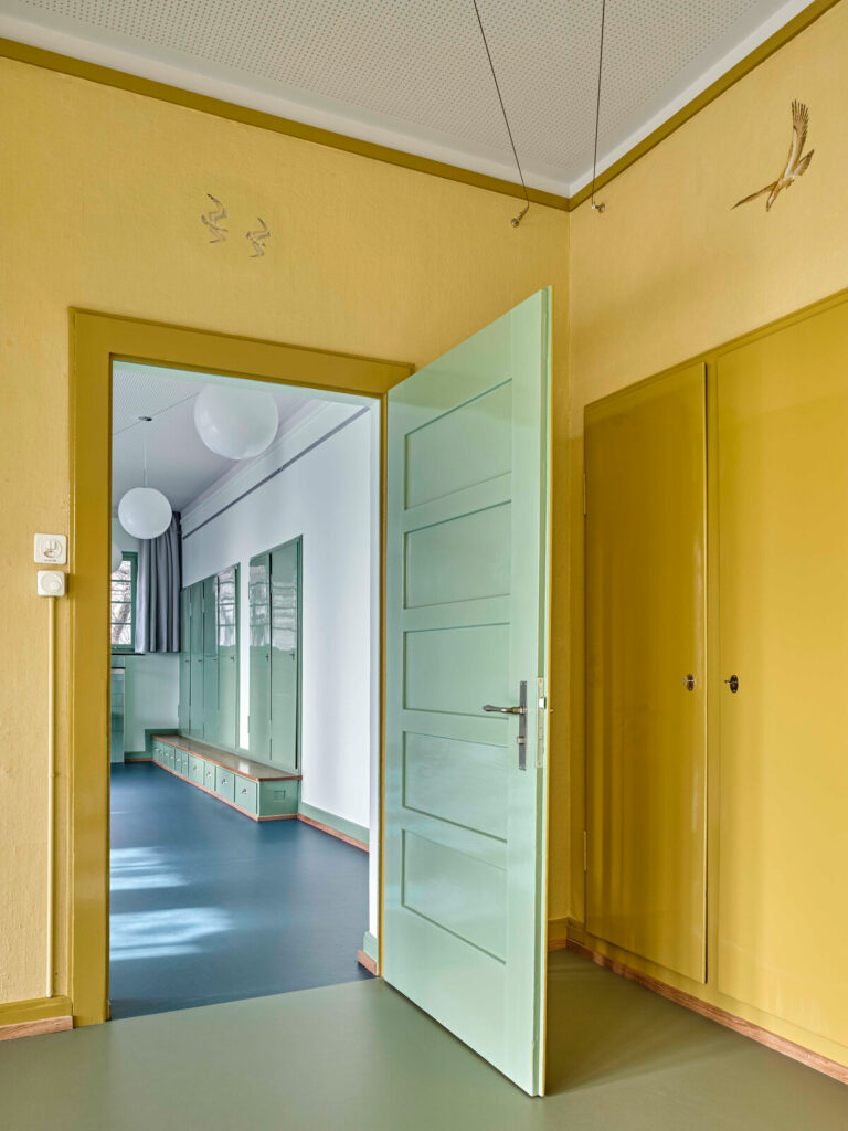 Geöffnete grüne Türe vor gelbem Wandschrank – beides restauriert – mit Blick auf die neuen Garderoben im Kindergarten Farenweg, umgesetzt durch die Schreinerei R. Brunner AG Zürich