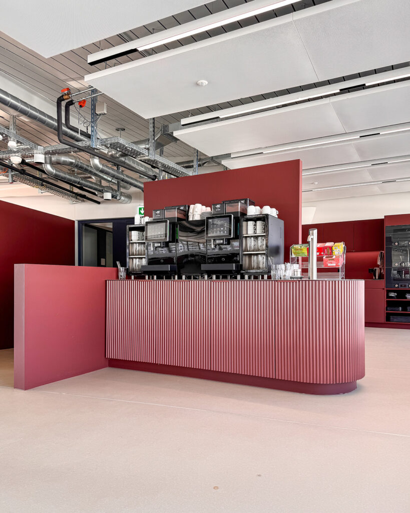 Freistehende Kaffeetheke mit weinroten Fronten und mehreren Kaffeemaschinen vor offenem Deckensystem