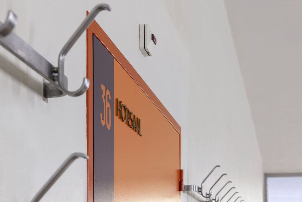Tür mit Raumbezeichnung „Hörsaal 36“ in Orange und Dunkelblau, flankiert von Wandhaken aus Edelstahl
