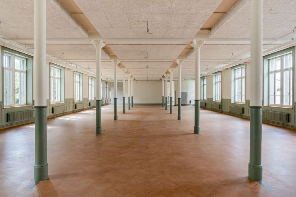 Eventsaal mit hohen Fenstern, grünen Wandtäfern und Stützenreihen