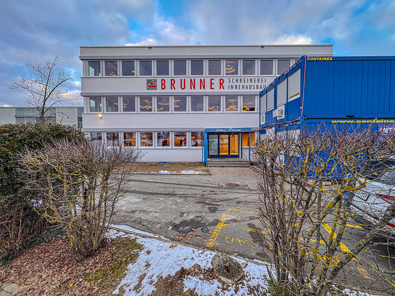Aussenansicht eines modernen Büro- und Werkstattgebäudes der Brunner Schreinerei Innenausbau mit grossem Logo an der Fassade, aufgenommen an einem Wintertag mit leichtem Schneefall auf dem Boden.