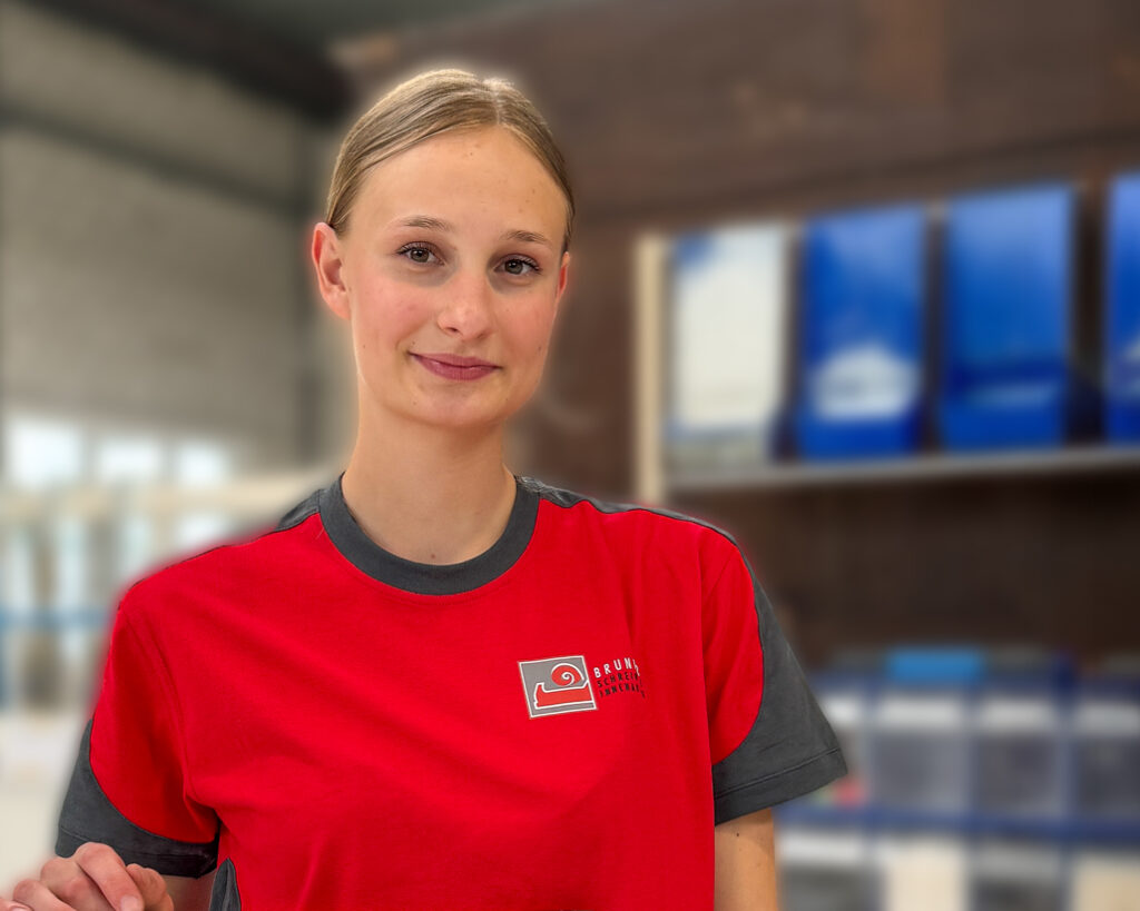 Eine junge Schreiner-Lernende im roten Betriebsshirt der Schreinerei R. Brunner AG lächelt in der Werkstatt und hält ein historisches Holzbauteil sowie eine Beisszange.