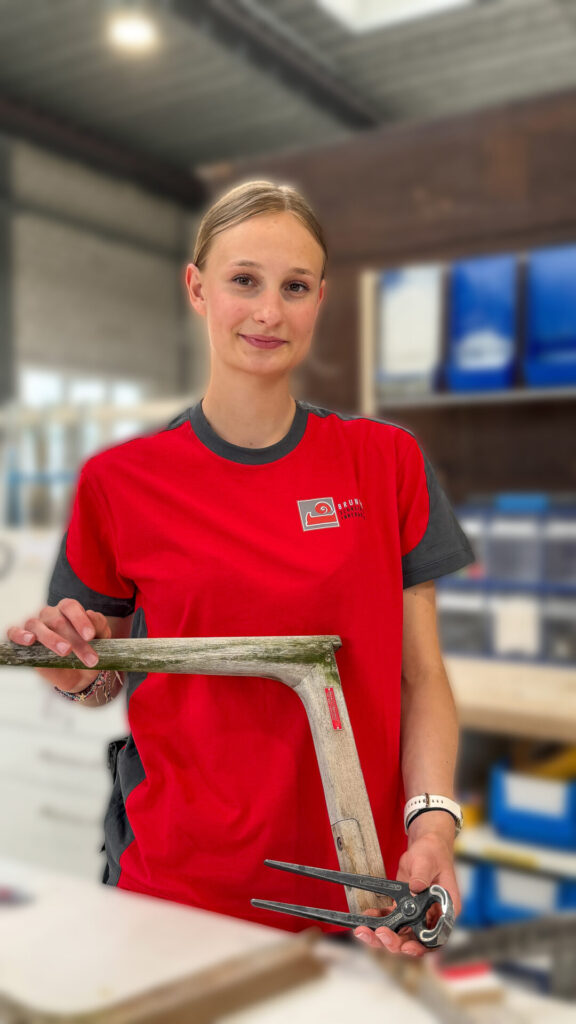 Eine junge Schreiner-Lernende im roten Betriebsshirt der Schreinerei R. Brunner AG lächelt in der Werkstatt und hält ein historisches Holzbauteil sowie eine Beisszange.