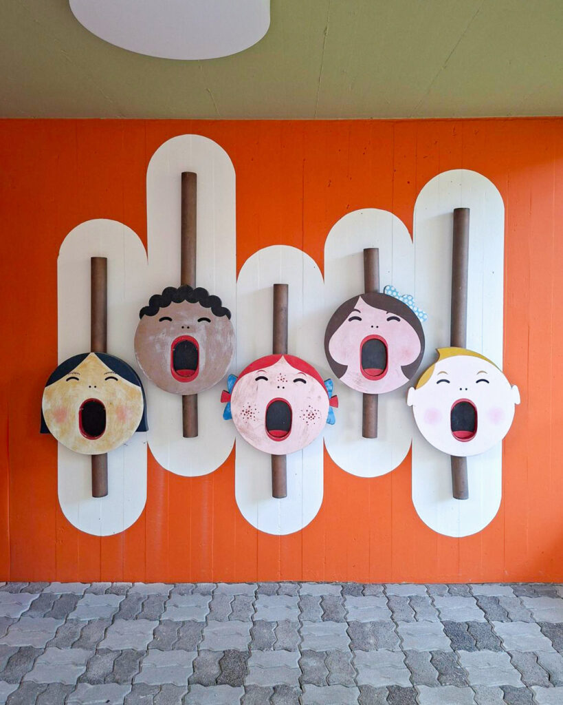 Fünf instand gesetzte Köpfe mit offenen Mündern vor orangefarbener Wand im Kindergarten Sihlweid in Zürich.